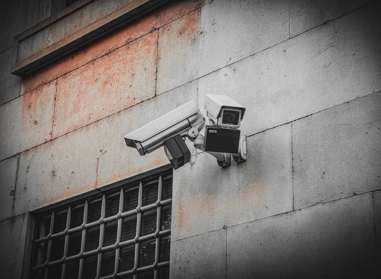 services-10 Two surveillance cameras mounted on an aged concrete wall beside a barred window, capturing urban security.